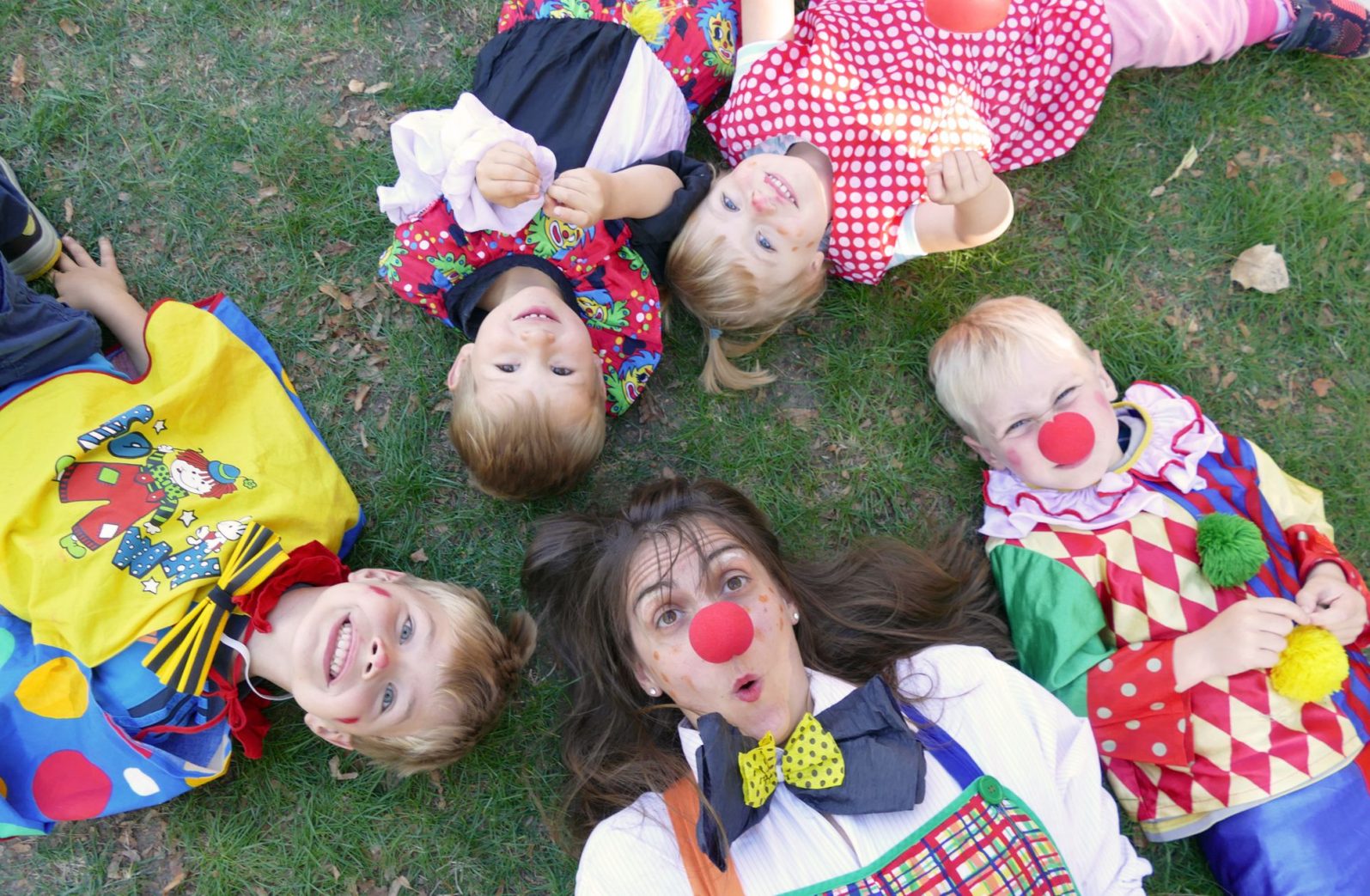 Kinder und Erzieherin liegen als Clowns verkleidet auf der Wiese